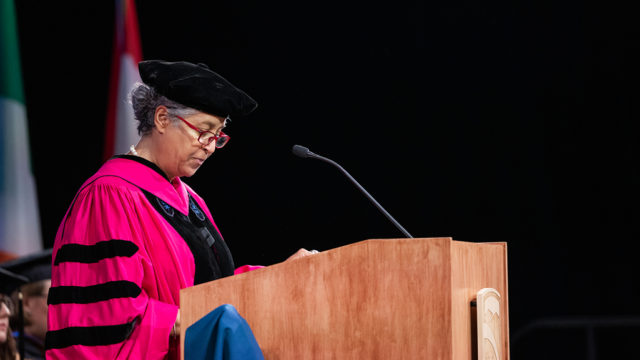 Monique Taylor, wearing her academic regalia, speaks into a microphone at a podium.