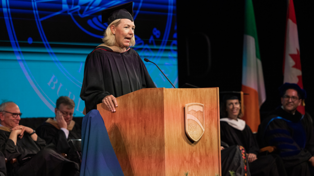 Judy O’Connell, wearing her academic regalia, speaks into a microphone at a podium.